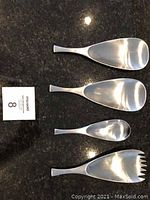 Four serving utensils (two large servers, one small spoon, one serving fork) laid out on dark surface