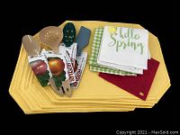 Set of 12 yellow vinyl placemats stacked with 4 new Hutzler utensils and 2 spring kitchen towels, plus red silicone pot holder on top.
