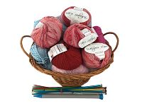 Wicker basket filled with six new balls of striped Loops & Threads Whisper Lace yarn in red, pink, and blue tones, along with a small assortment of colorful knitting needles laid in front.