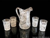 Set of one crystal pitcher with four matching glasses displayed on black reflective surface showing intricate geometric and pinwheel cut patterns.