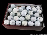 Top-down photo showing a box filled with approximately 84 white golf balls with visible logos and wear marks.
