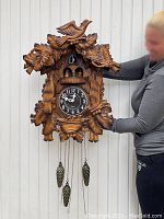 Full front view of the large Black Forest style cuckoo clock hanging, showing intricate carved leaf and bird motifs, clock face, dancers on a rotating disk, and hanging ornamental pine cone-shaped weights.