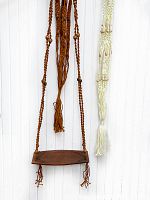 Both macramé hanging teak shelf with carved details and cream-colored macramé plant hanger with wooden beads hanging against white wall.