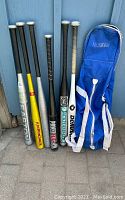 Photo of six aluminum softball bats and one blue carry bag against a blue wall.