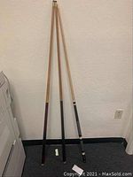 Three full length pool cues standing upright against a wall, showing condition and design of grips and shafts.