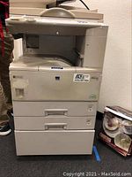 Front and side view of Ricoh Aficio 220 printer on rollers placed in a room, showing paper trays and control panel, item condition and size.