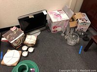Photo showing all items in the lot: TV, dinnerware set, champagne chiller with glasses, boxed hair dryer and lantern, and miscellaneous items on floor.