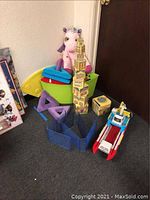Wide shot showing Paw Patrol cargo ship, foam building blocks, plush unicorn, cardboard dollhouse blocks stacked tall, and blue plastic blocks arranged in a circle on carpeted floor.