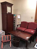 Faux grandfather clock bookshelf beside a two-seater red upholstered wooden chair, rectangular wooden coffee table, wooden magazine rack, and a woven waste bin on floor.