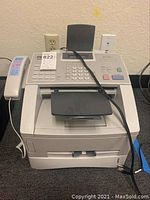 Image showing front view of white business class laser fax machine with keypad, display screen, and handset, with cables attached.