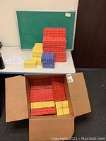 Photo showing cardboard building blocks in red, yellow, and blue stacked, with a green chalkboard behind them on a table.