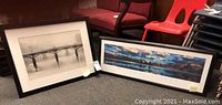 Two framed prints placed on floor near red chairs, showing black and white bridge print and panoramic color landscape print.