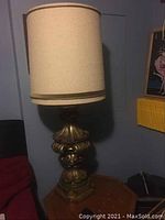 Tall table lamp with visible beige fabric drum shade and ornate carved Italian style gold and green toned base on wooden side table.
