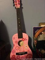 Front view of child's pink acoustic guitar with floral design and black fretboard against a gray wall.