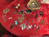 All earrings arranged on a red surface with heart-shaped tan paper box visible. Variety of styles, colors and materials shown.