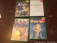 Four Xbox game cases on a wooden surface showing titles and covers for X-Men Legends, SSX 3 Platinum Hits, an obscured game, and Shark Tale.