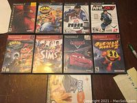 Top-down view of nine PlayStation 2 game cases arranged on a wooden surface, showing cover artwork of various game titles including Crash Bandicoot, NHL 2004, The Sims, Cars, and Pac-Man World 2.