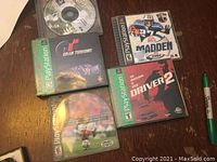 Five PlayStation 1 game discs and cases placed on a wooden surface. Titles visible include Madden 2001, Gran Turismo, Driver 2, and two football games. A green marker is placed beside the games for scale.