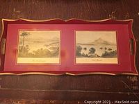 Top view of rectangular red and gold serving tray displaying two vintage landscape prints under glass with side handles.
