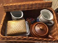 Basket containing decorative items including ceramic teapot, two human-shaped ceramic jugs, metal ice cubes, floral box, and plastic container