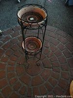 Full view of black metal plant stand showing two terracotta pots on round shelves with curved legs on a brick floor.