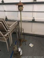 Full view of tall cast iron candle holder with conical roof and wire plant stand beside it, stacked chairs in background