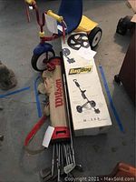 Photo of the BagBoy golf push cart in box with Wilson golf bag and golf clubs laid alongside, children's tricycle partially visible.