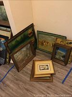 Photo showing multiple framed Asian wall art paintings resting against walls and floor, varying in size and style featuring traditional scenes and figures.