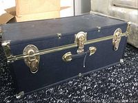 Front view of black steamer trunk showing brass lock, side latches, and handle. Surface has scratches and marks.