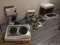 Wide shot showing two white Jenn Air stovetops with two black coil burners each, cardboard boxes, a wire rack, trays, bowls, and pepper hanging kitchen decor on greyish wooden floor.