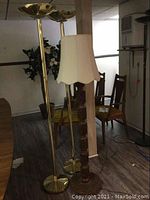 Full view of two gold-colored halogen floor lamps alongside one carved wood floor lamp with fabric lampshade in a room with wooden flooring and chairs in background.