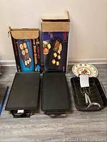 Photo of the two boxed electric griddles laid flat on floor along with the roaster pan with rack and the round platter.