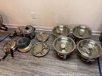 Collection of metal kitchen items including slow cooker, pressure cookers, metal divided plates, and sterno pots arranged on the floor.