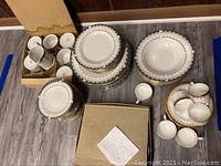 Full view of stacked plates, soup bowls, cups and saucers all with green and gold detailing in EXCEL Ardsley pattern, along with boxed Hitkari cups and saucers.