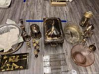 Overview of all silver plate and glass serving items on floor including teapot, pitcher, platters, cordial cups, candle holder, chafing dish and glass bowl with silver base.