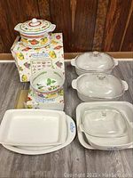 Photo showing 3 white Corning Ware cornflower serving platters, 1 Corning Ware casserole pan with lid, 2 white Corning Ware F14B casserole pans with lids, and 2 Fresh N Fruity bowls, one with lid on boxes.