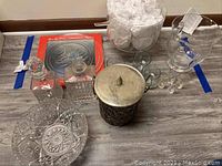 Wide view of the lot showing cut glass bowl, Christmas tree platter in box, punch bowl filled with wrapped items, metal ice bucket, shot glasses, whiskey glasses, and two decanters on a table.