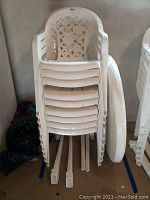 Stack of white plastic patio chairs with lattice pattern backrests and slatted seats, dusty and dirty. Chairs are stacked with legs visible.