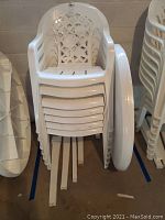 Stack of 7 white plastic patio chairs with lattice pattern on the back, armrests, and slotted seats. Stacked chairs shown from the front with visible dirt and dust.