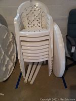 Stack of eight white plastic patio chairs with lattice backs and armrests, dusty and in need of cleaning. White round plastic table top stored vertically next to the chairs.