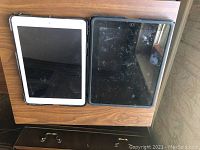 Top-down view of two iPads placed side by side on a wooden table surface. The left iPad has a white bezel, the right iPad has a black bezel with visible smudges on screen.