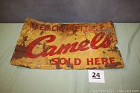 Front view of rectangular metal sign with 'Camels Sold Here' and faded 'Need Cigarettes?' text, showing rust and paint wear.