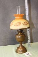 Full view of the complete oil lamp showing brass base, burner, floral dome shade with ruffled edge, and clear chimney against a wall with a ruler for height reference.