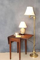 Overall view showing the drop leaf table with small table lamp (ceramic base, floral) and tall floor lamp (brass tone), also showing cup, saucer and needlework doily on table