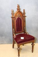 Front view of the carved wooden church chair showing pointed arch back with red velvet upholstery and decorative brass tacks.