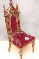 Full front and side view of the ornate wooden church chair showing red velvet upholstery on seat and backrest with detailed carving and finials.
