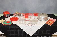 Wide view of lot including red glass storage container, glass butter keeper, red plastic container, three red tin trays, a metal dome cover, red kitchen scale, four red divided plates, two pot holders, three glasses, and embroidered linens