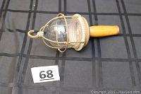 Industrial light with protective metal cage around glass globe, wooden handle and hanging hook, resting on black surface with lot number card 68.