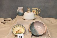 Full set of pottery items on display fabric background: cheese dome with mouse handle, a dog mug signed Nancy Anderson, a small turtle mug, a bunny bowl, and a blue and cream bowl with floral pattern and handles.