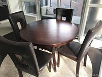 Photo of round solid wood dining table with four matching dark faux leather upholstered chairs set on a carpet in a bright room with large windows.